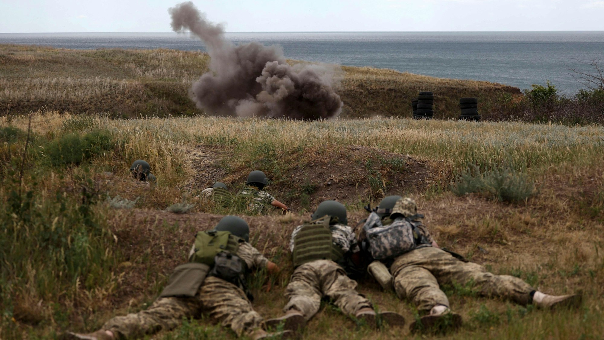 Soldatinnen und Soldaten liegen bei einer Militärübung in Odesa auf dem Boden, im Hintergrund eine Explosion