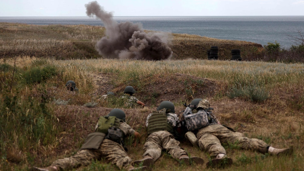Soldatinnen und Soldaten liegen bei einer Militärübung in Odesa auf dem Boden, im Hintergrund eine Explosion