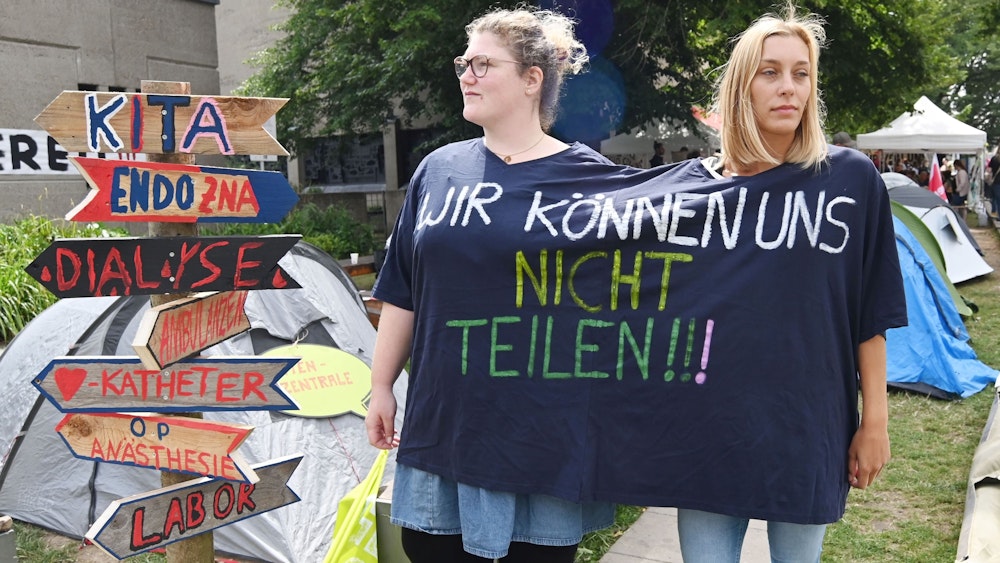 Zwei Streikende vor der Kölner Uniklinik in einem gemeinsam getragenen T-Shirt mit der Aufschrift "Wir können uns nicht teilen!"