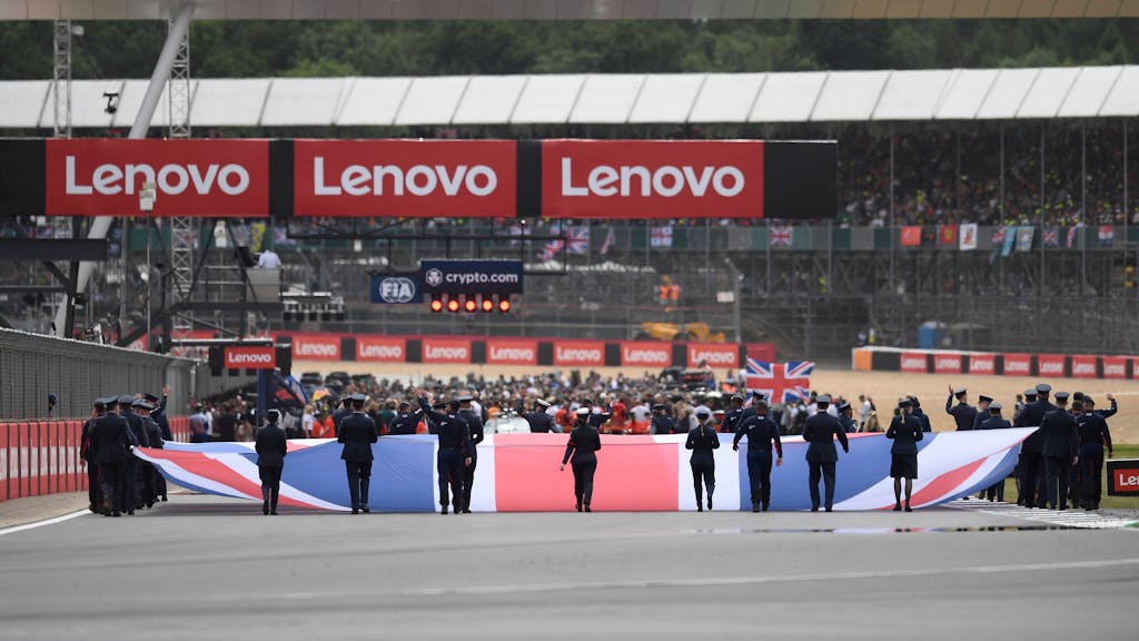 Auf der Start- und Zielgeraden hissen mehrere Personen die Flagge Großbritanniens – im Hintergrund bereiten sich die Teams auf das anstehende Rennen vor.