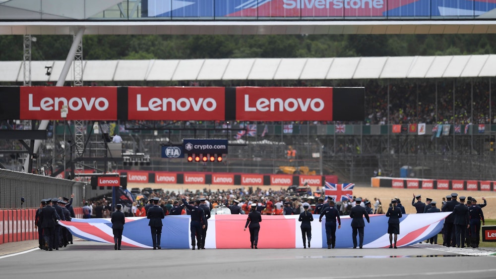 Auf der Start- und Zielgeraden hissen mehrere Personen die Flagge Großbritanniens – im Hintergrund bereiten sich die Teams auf das anstehende Rennen vor.