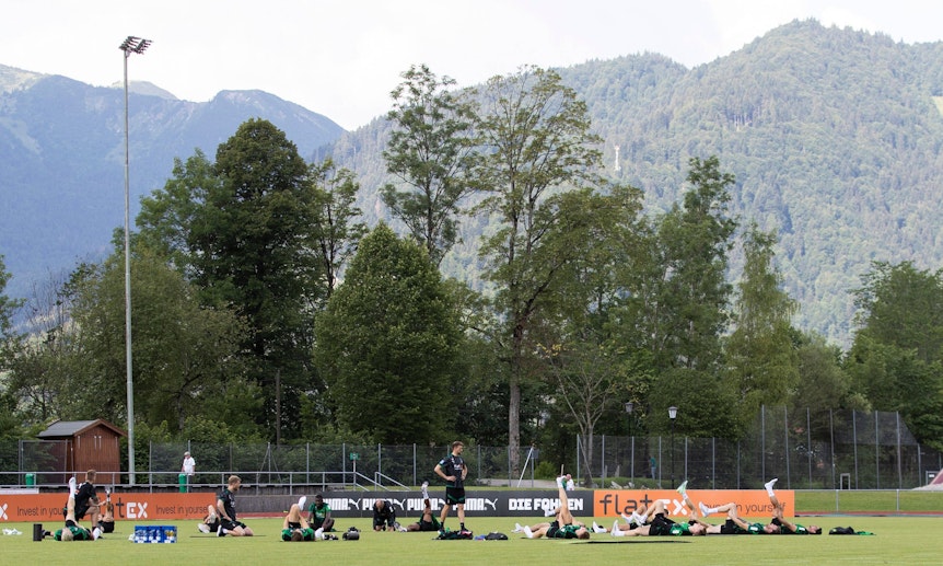 Die Gladbach-Profis im Trainingslager (6. Juli 2022) am Tegernsee in Rottach-Egern. Im Hintergrund sind die Bayerischen Alpen zu sehen. Die Spieler liegen auf dem Rasen und machen Stretching.