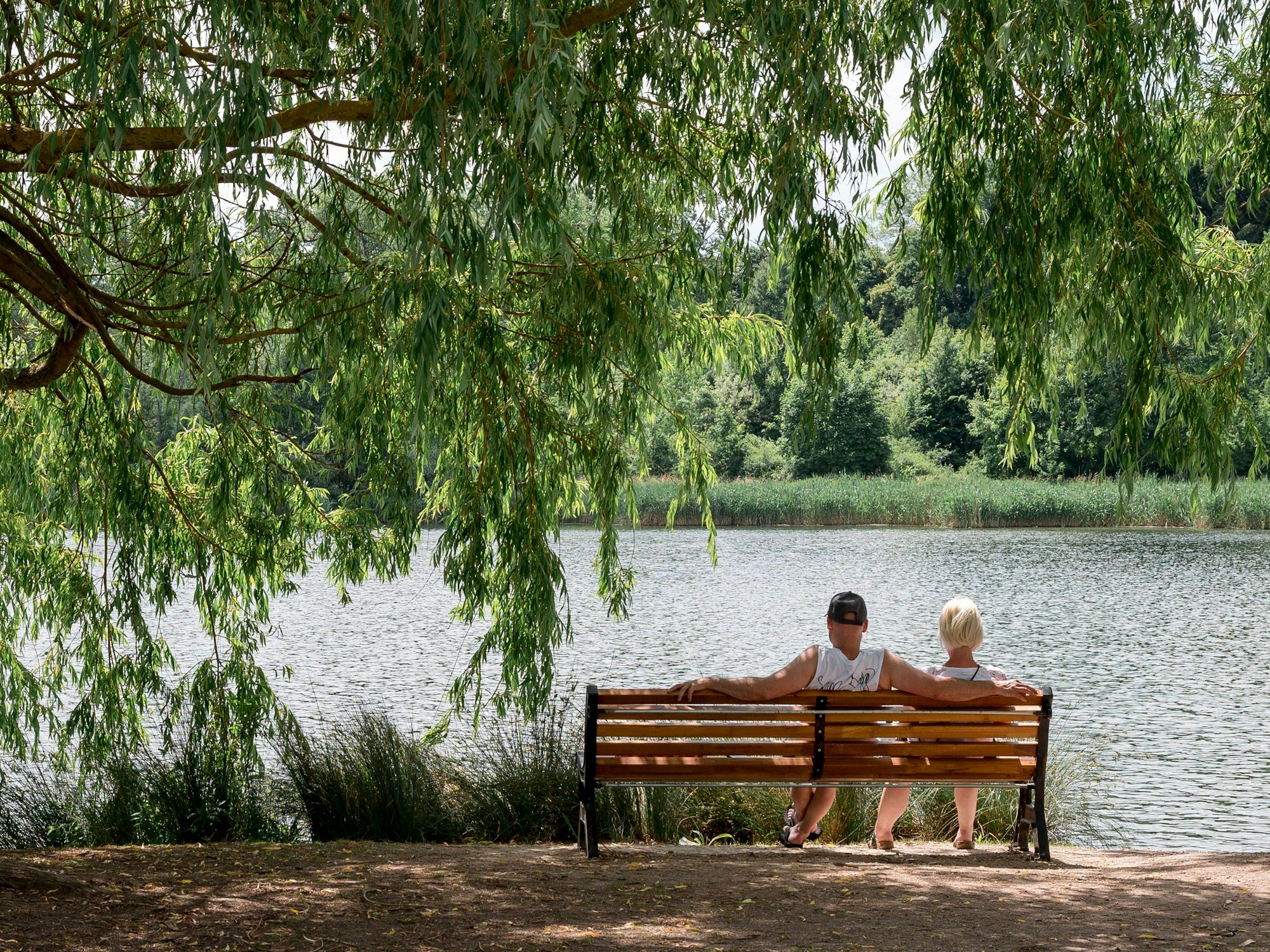 Ein Paar sitzt auf einer Bank in der Sonne an einem See.
