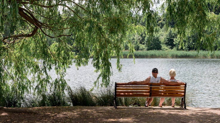 Ein Paar sitzt auf einer Bank in der Sonne an einem See.