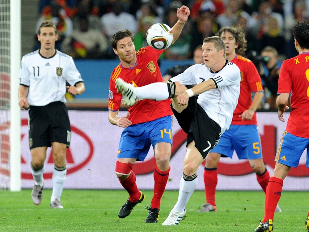Deutschlands Bastian Schweinsteiger schlägt vor den Augen seines spanischen Gegenspielers Xabi Alonso den Ball weg. Im Hintergrund sind Miroslav Klose und Carles Puyol zu sehen.
