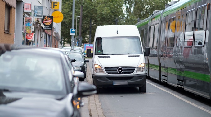 Ein Lieferwagen steht auf einer Straße neben parkenden Autos, daneben fährt eine Straßenbahn entlang.