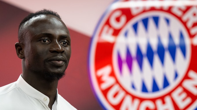 Sadio Mané posiert im weißen Hemd, hinter ihm ist das Wappen des FC Bayern zu sehen.