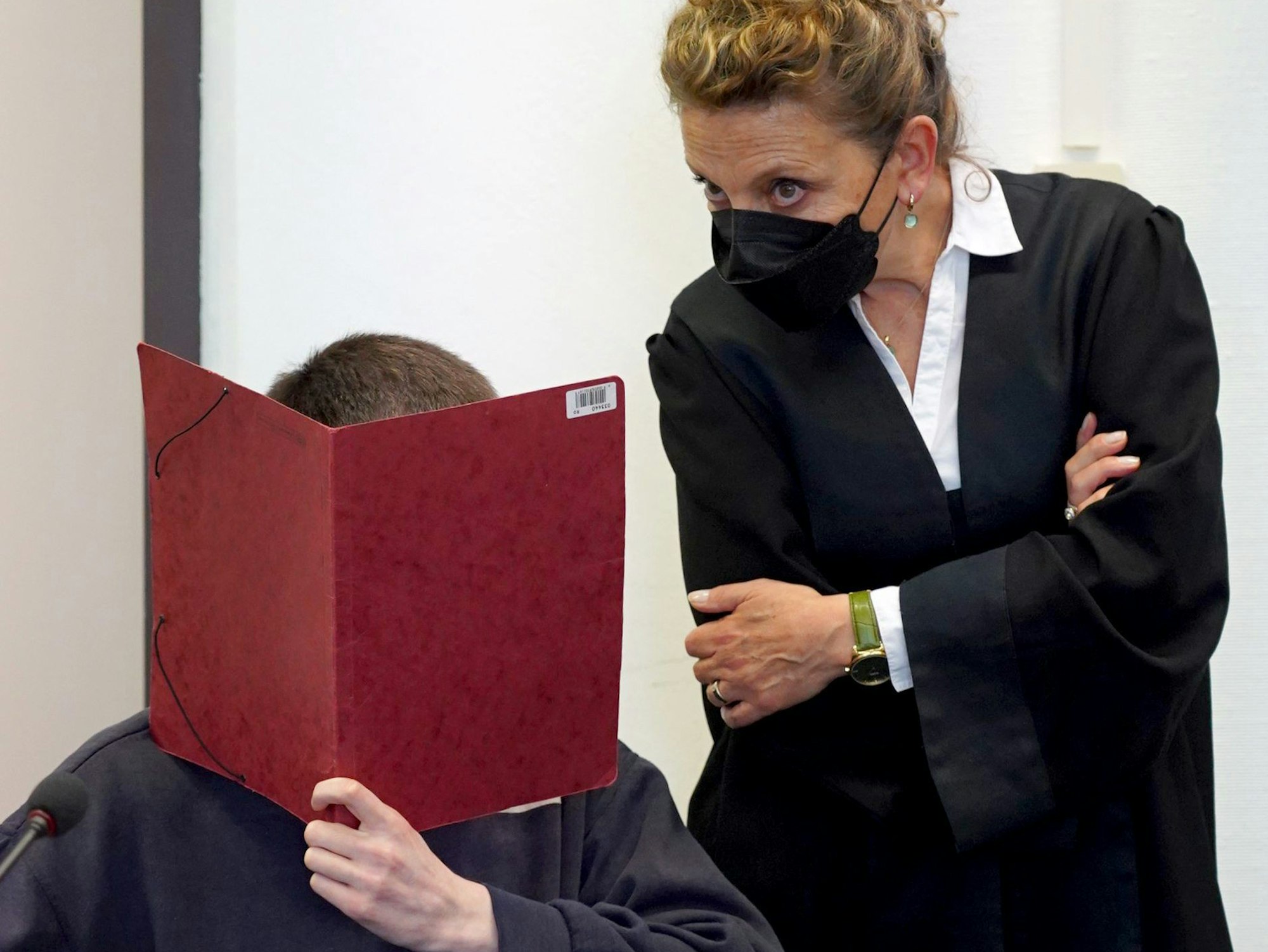 Ein Mann hält sich in einem Gerichtssaal eine rote Mappe vor das Gesicht.