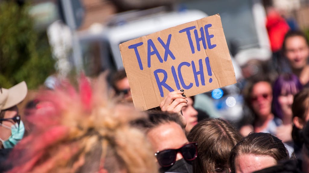 Teilnehmer einer Demonstrantion des Bündnisses „Wer hat, der gibt“ halten ein Schild mit der Aufschrift „Tax The Rich!“.