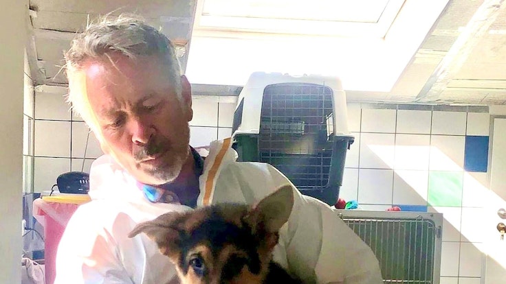 A man holds German shepherd puppy Poldi in his arms.  In the background are animal boxes in a tiled room.