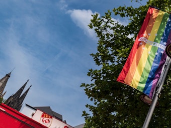 Eine LGBTIQ+-Flagge hängt an einer Straßenlaterne, im Hintergrund die Spitzen des Kölner Doms.