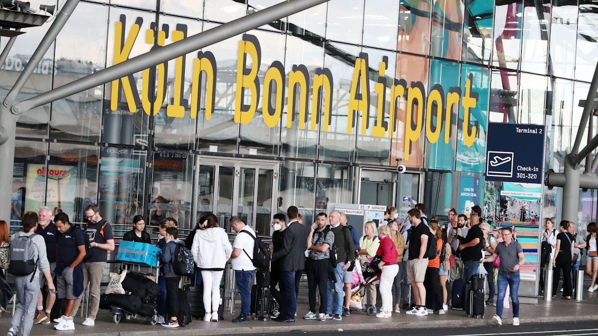 Fluggäste stehen vor einem Gebäude mit der Aufschrift "Köln Bonn Airport" in der Warteschlange.&nbsp;