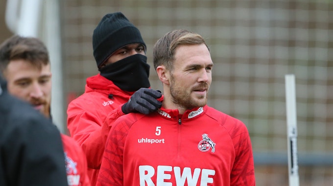 Rafael Czichos vom 1. FC Köln steht beim Training auf dem Platz, Anthony Modeste, der sich in Schal und Mütze gehüllt hat, steht hinter ihm.