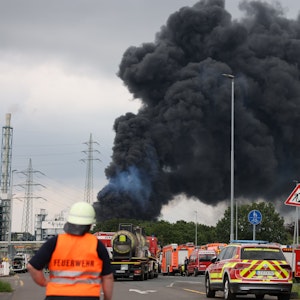 Einsatzfahrzeuge der Feuerwehr stehen unweit einer Zufahrt zum Chemiepark, über dem eine dunkle Rauchwolke aufsteigt. Am 27. Juli 2021 war es in Leverkusen zu einer verheerenden Explosion gekommen. Sieben Mitarbeiter starben, viele wurden verletzt.