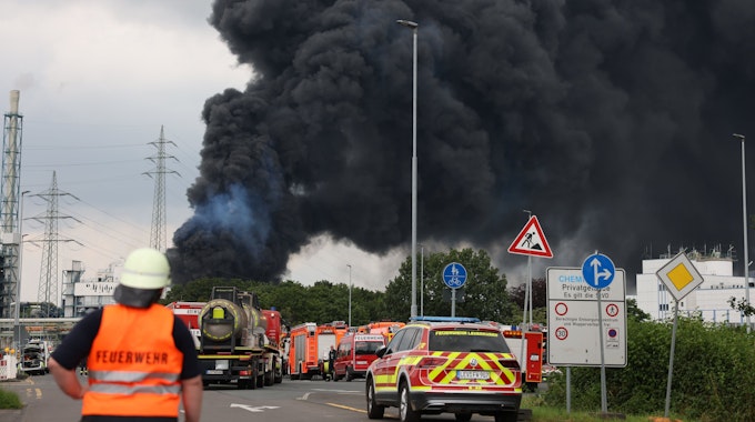 Einsatzfahrzeuge der Feuerwehr stehen unweit einer Zufahrt zum Chemiepark, über dem eine dunkle Rauchwolke aufsteigt. Am 27. Juli 2021 war es in Leverkusen zu einer verheerenden Explosion gekommen. Sieben Mitarbeiter starben, viele wurden verletzt.