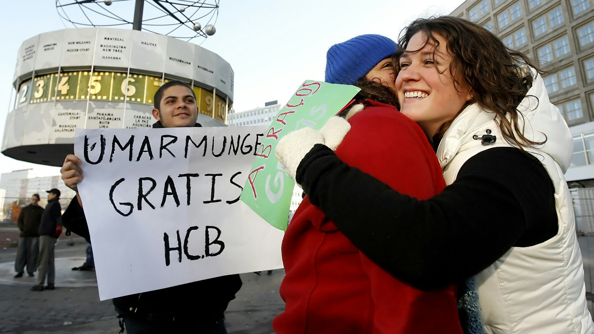 Menschen geben sich auf dem Alexanderplatz in Berlin eine Umarmung, ein weiterer hält ein Schild mit dem Text „Umarmung gratis“ hoch.