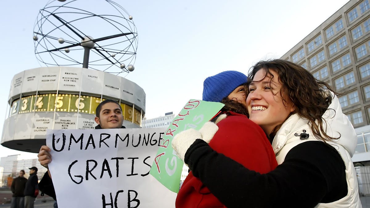 Menschen geben sich auf dem Alexanderplatz in Berlin eine Umarmung, ein weiterer hält ein Schild mit dem Text „Umarmung gratis“ hoch.