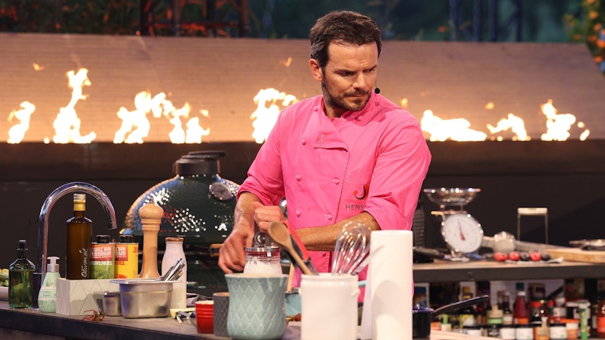 Steffen Henssler, steht samt rosa Koch-Bekleidung in der Küche.