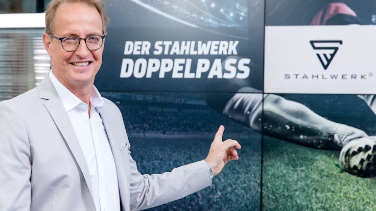 Florian König zeigt auf das Logo des Fußball-Talks Doppelpass. Er wird den Doppelpass auch in der Saison 2022/2023 moderieren.