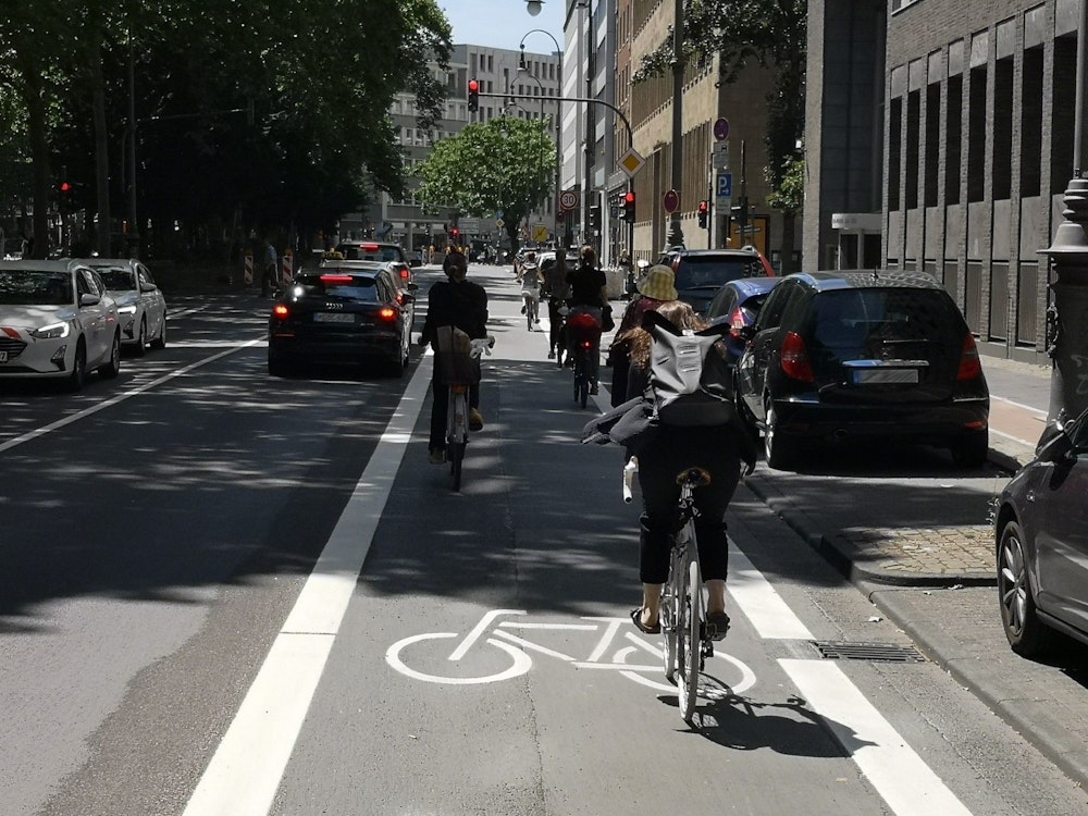 Foto des neuen Radfahrstreifens auf dem Kaiser-Wilhelm-Ring. Zu sehen sind einige Radfahrende, die den Radfahrstreifen befahren, sowie parkende und fahrende Autos.