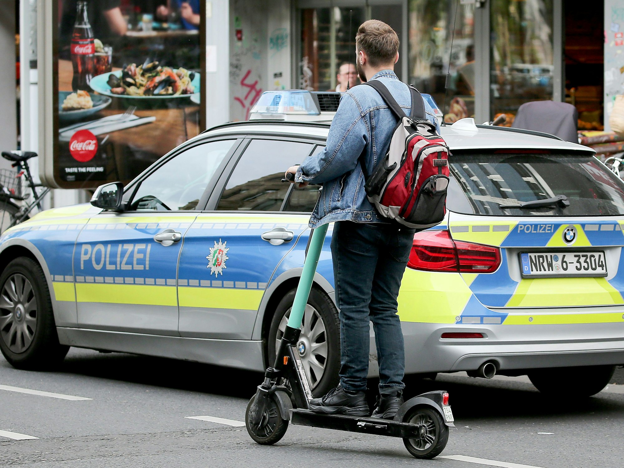 Mit einem Elektro-Tretroller fährt ein Mann an einem Streifenwagen der Polizei vorbei.
