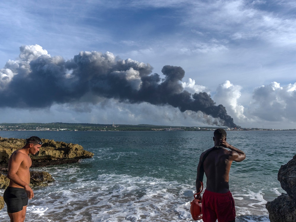 Sonntag, 7. August: Zwei Männer beobachten, wie eine riesige Rauchwolke vom Supertankerstützpunkt aufsteigt, während Feuerwehrleute daran arbeiten, ein Feuer zu löschen, das in der Nacht zuvor während eines Gewitters ausgebrochen war. Nach einer Reihe von Explosionen in einem Treibstofflager im Norden von Kuba kämpfen die Einsatzkräfte immer noch gegen die Flammen.
