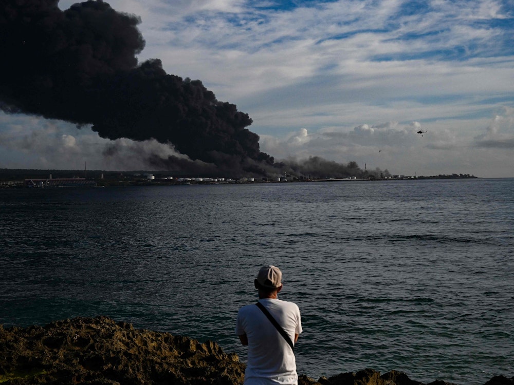 TOPSHOT - A man looks at black smoke rising from a massive fire at a fuel depot sparked by a lightning strike in Matanzas, Cuba, on August 8, 2022. - A second oil tanker collapsed at midnight Sunday in Matanzas, in western Cuba, where firefighters have been battling a huge blaze for two days, which has left one dead and 16 missing, local authorities reported. (Photo by YAMIL LAGE / AFP)