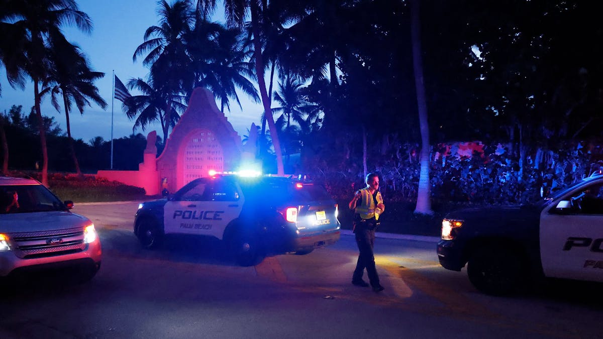 Das undatierte Symbolfoto zeigt drei US-amerikanische Polizeiwagen mit Blaulicht und einen uniformierten Polizisten vor einem Tor. Überall stehen Palmen.