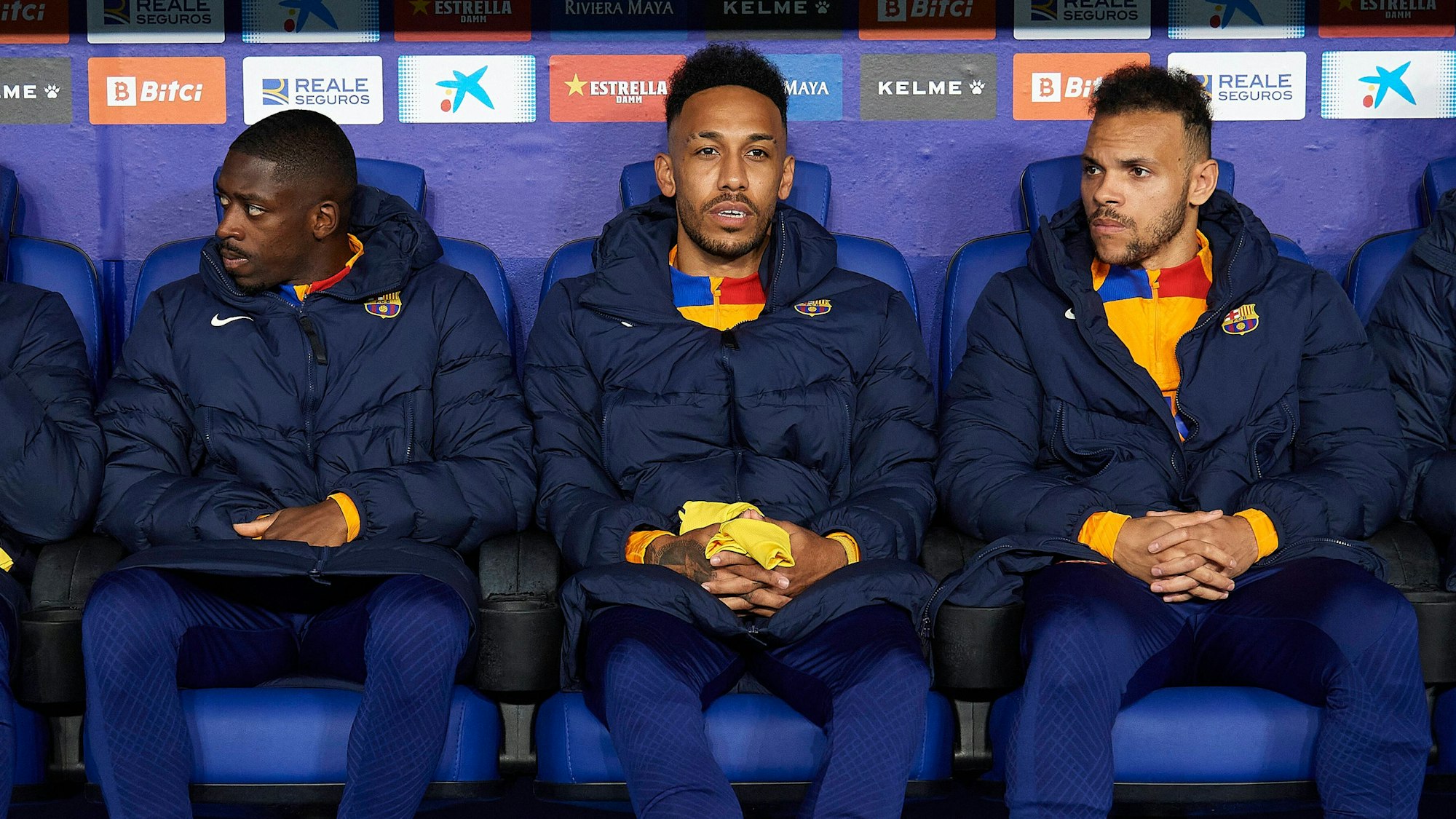 Ousmane Dembele, Pierre-Emerick Aubameyang und Martin Braithwaite (von l. nach r.) vom FC Barcelona sitzen mit wärmenden Jacken auf der Bank.