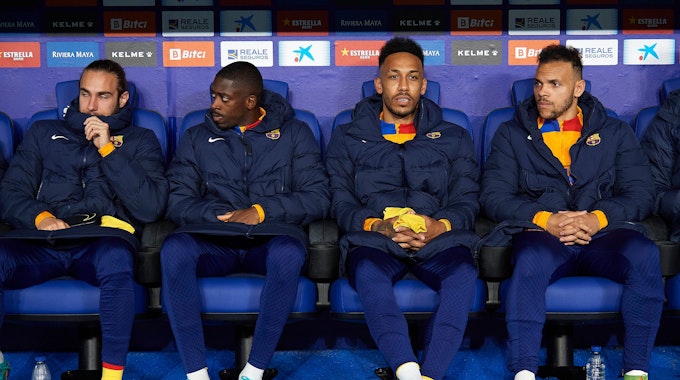 Ousmane Dembele, Pierre-Emerick Aubameyang und Martin Braithwaite (von l. nach r.) vom FC Barcelona sitzen mit wärmenden Jacken auf der Bank.