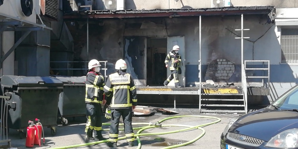 Ein Feuerwehrmann kommt aus einem Gebäude, in dem es gebrannt hat, zwei Kollegen stehen davor, zu ihren Füßen Löschschläuche.