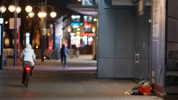 Ein Mensch liegt in der Innenstadt in einem Schlafsack im Eingang zu einem Geschäft. Die FDP will einen Antrag für ein Pilotprojekt für Köln stellen – eine Alarmanlage für Obdachlose soll her.