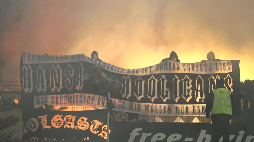 Hansa-Rostock-Fans zeigen auf dem Zaun des Gästeblocks ein Banner, im Hintergrund brennt Pyrotechnik.