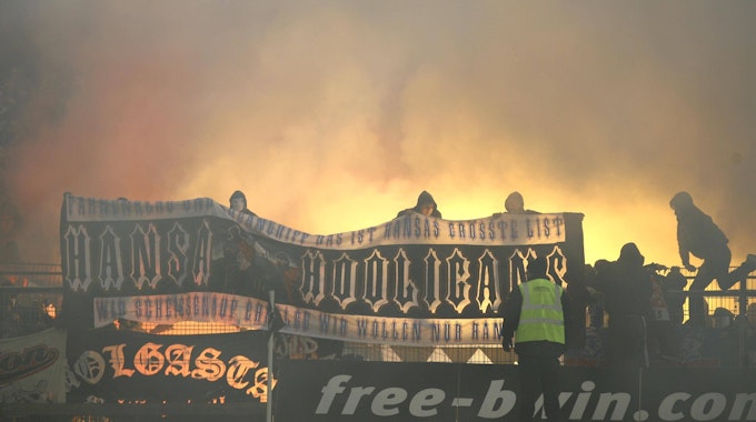 Hansa-Rostock-Fans zeigen auf dem Zaun des Gästeblocks ein Banner, im Hintergrund brennt Pyrotechnik.