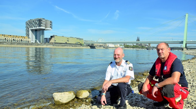 Zwei Kölner Experten hocken direkt am Rheinufer und blicken in die Kamera. Die Gefahren am Rhein sind größer als je zuvor, sagen sie.