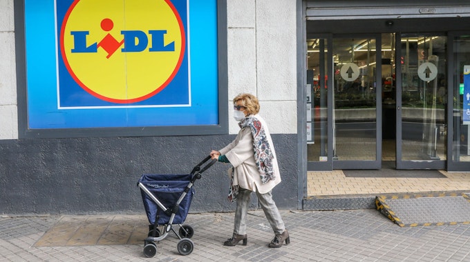 Eine Frau mit Rollator trägt einen Mund-Nasen-Schutz und passiert ein Einkaufsladen von Lidl.