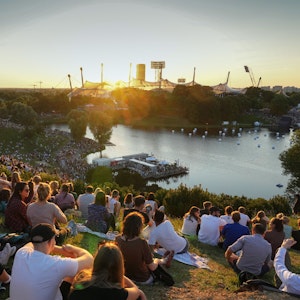 Menschen sitzen auf dem Münchner Olympiaberg und schauen auf den See vor dem Olympiastadion.