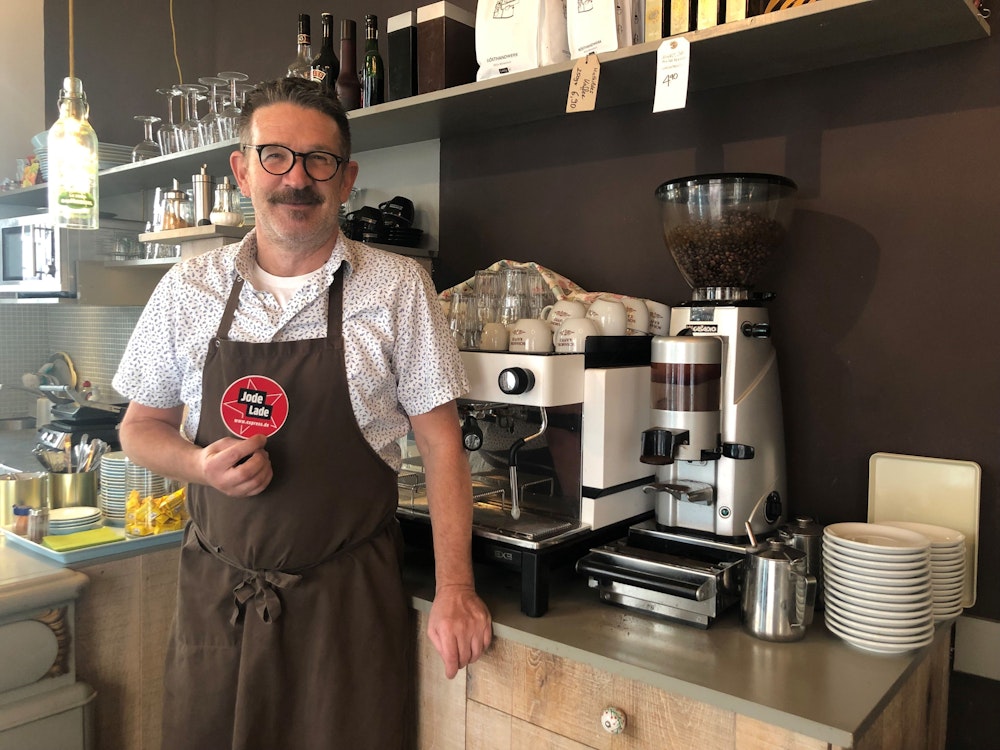 Thomas Kastner steht vor der Kaffeemaschine in seinem Café und hält den „Jode Lade“-Sticker in die Kamera.