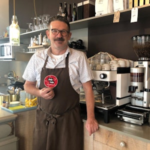 Thomas Kastner steht vor der Kaffeemaschine in seinem Café und hält den „Jode Lade“-Sticker in die Kamera.