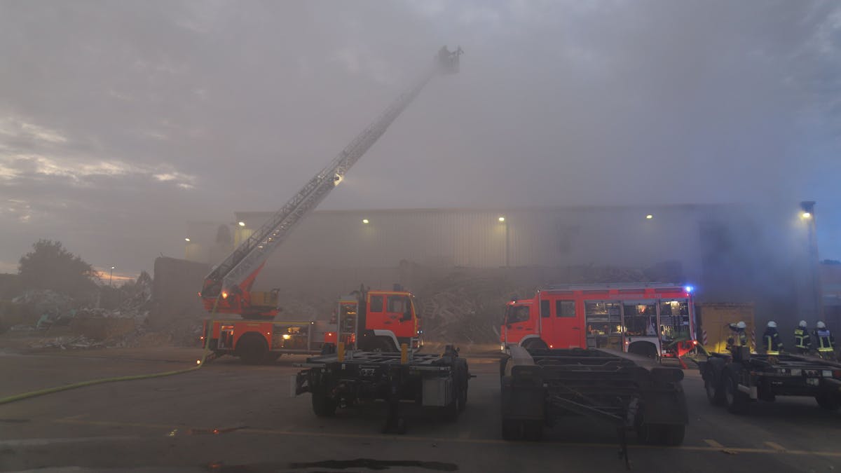Rauch dringt aus einer Halle, vor der zwei Löschfahrzeuge der Feuerwehr stehen. Es hat die Drehleiter ausgefahren.&nbsp;