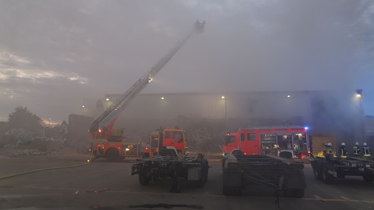 Rauch dringt aus einer Halle, vor der zwei Löschfahrzeuge der Feuerwehr stehen. Es hat die Drehleiter ausgefahren.