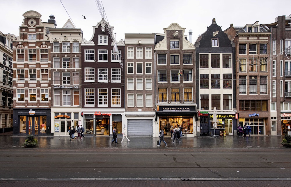 Das Bild zeigt eine Häuserreihe in den Niederlanden mit Ladenlokalen im Erdgeschoss. Auf dem Bürgersteig laufen Passanten und Passantinnen.