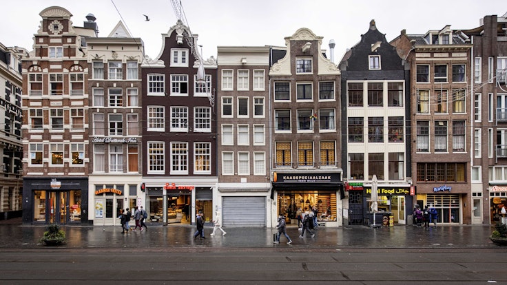 Das Bild zeigt eine Häuserreihe in den Niederlanden mit Ladenlokalen im Erdgeschoss. Auf dem Bürgersteig laufen Passanten und Passantinnen.