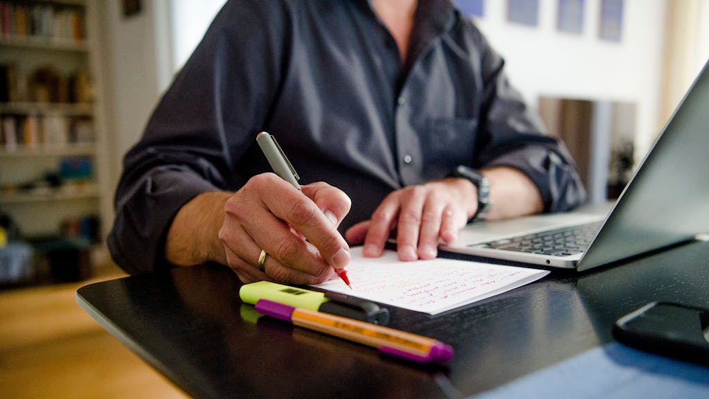 Ein Mann sitzt zuhause an einem Esstisch. Er schreibt mit einem Stift auf ein Blatt Papier neben einem Laptop
