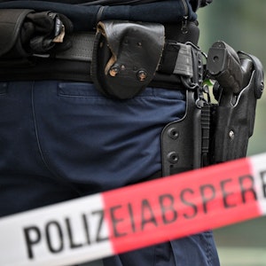 Das undatierte Symbolfoto zeigt ein rot-weißes Flatterband mit der Aufschrift „Polizeiabsperrung“. Dahinter steht eine Person mit einer Waffe im Holster.