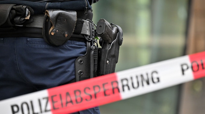 Das undatierte Symbolfoto zeigt ein rot-weißes Flatterband mit der Aufschrift „Polizeiabsperrung“. Dahinter steht eine Person mit einer Waffe im Holster.