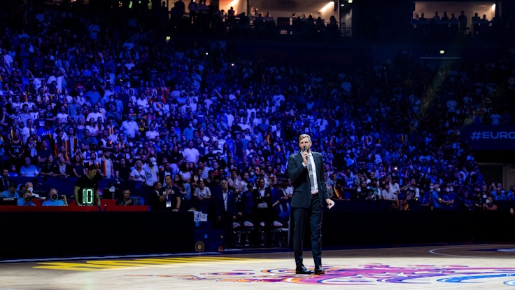 Dirk Nowitzki steht in der abgedunkelten Lanxess-Arena in Köln und hält mit Mikrofon eine Rede.