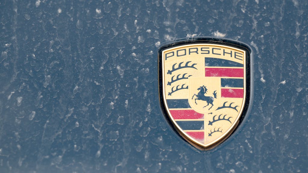 Ein Logo des Fahrzeugbauers Porsche ist an einer mit Wüstenstaub bedeckten Motohaube eines Sportwagens angebracht. 