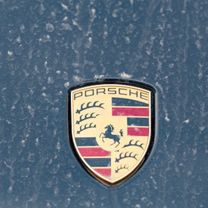 Ein Logo des Fahrzeugbauers Porsche ist an einer mit Wüstenstaub bedeckten Motohaube eines Sportwagens angebracht.