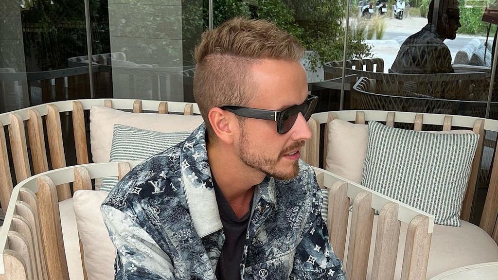 Influencer Julian Claßen, hier auf einem Instagram-Bild von Juni 2022, macht mit seiner neuen Freundin Tanja Makaric den nächsten Schritt. Julian sitzt in einem Korbstuhl, er trägt ein schwarz-weiß gemustertes Hemd und eine schwarze Sonnenbrille.